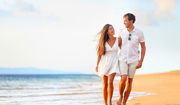 Beach couple walking on romantic travel honeymoon vacation summer holidays romance. Young happy lovers, Asian woman and Caucasian man holding hands embracing outdoors.
