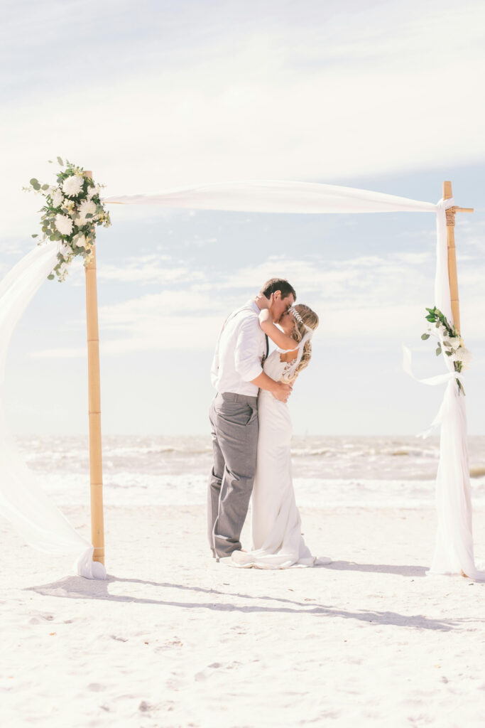 Bride and groom sharing a kiss under a simple beach wedding arch decorated with white flowers and fabric, perfect for a destination wedding.