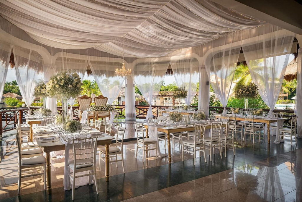Elegant covered wedding reception with white draping, chandeliers, and floral centerpieces overlooking a tropical resort setting.