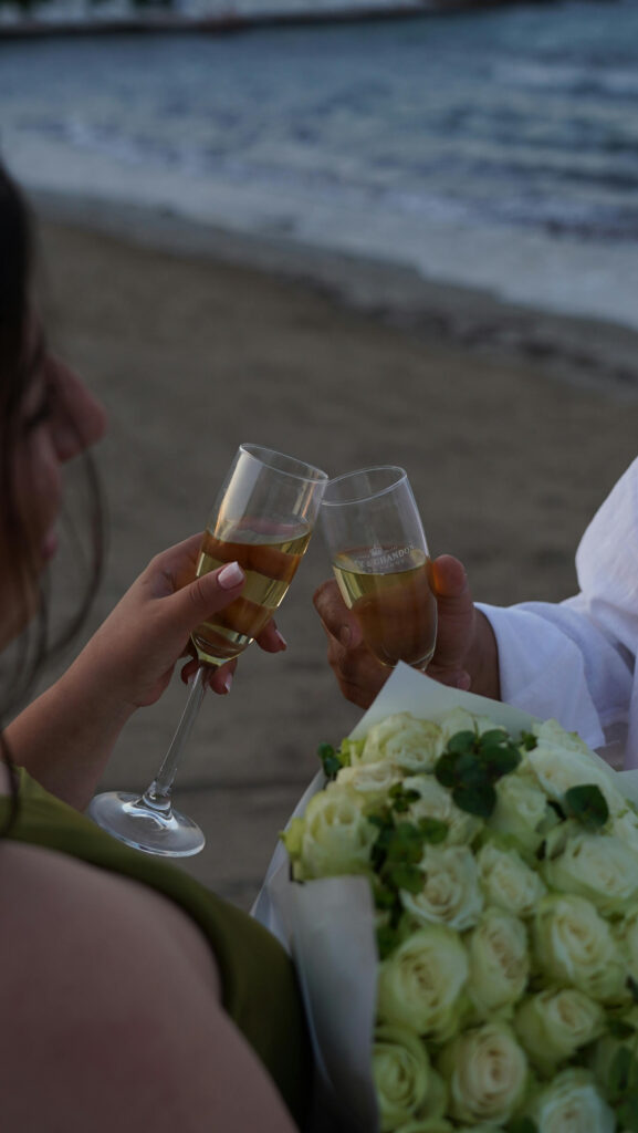 Close-up of couple clinking champagne glasses on the beach with a bouquet of white roses.