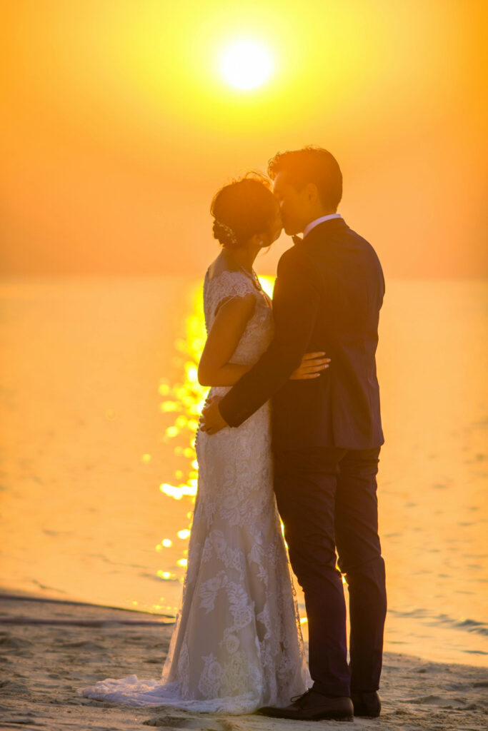 Bride and groom share a romantic kiss at sunset with golden light reflecting on the ocean.